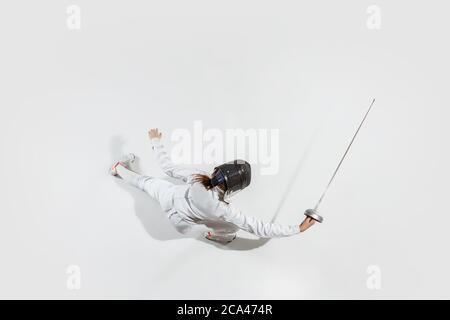 D'abord. Jeune fille en costume d'escrime avec épée à la main sur fond blanc. Vue de dessus. Jeune femme modèle pratique et formation en mouvement, action. CopySpace. Sport, jeunesse, mode de vie sain. Banque D'Images