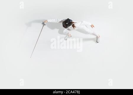 D'abord. Jeune fille en costume d'escrime avec épée à la main sur fond blanc. Vue de dessus. Jeune femme modèle pratique et formation en mouvement, action. CopySpace. Sport, jeunesse, mode de vie sain. Banque D'Images