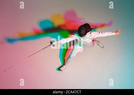 Couleur. Jeune fille en costume d'escrime avec une épée à la main sur fond de dégradé à la lumière du néon. Vue de dessus. Jeune modèle pratiquant, formation en mouvement, action. CopySpace. Sport, jeunesse, mode de vie sain. Banque D'Images