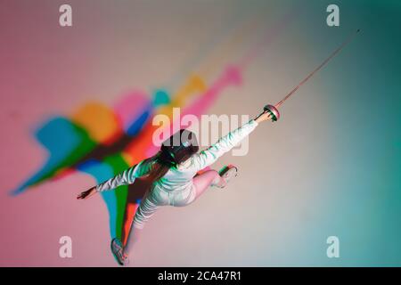 Couleur. Jeune fille en costume d'escrime avec une épée à la main sur fond de dégradé à la lumière du néon. Vue de dessus. Jeune modèle pratiquant, formation en mouvement, action. CopySpace. Sport, jeunesse, mode de vie sain. Banque D'Images