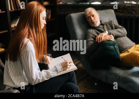 Jeune femme sérieuse à cheveux rouges psychologue prenant des notes dans le carnet pendant la séance de psychothérapie. Banque D'Images