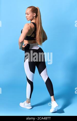 Vue arrière d'une jeune femme mince et en bonne santé dans les vêtements de sport de mode. Image pleine longueur du modèle femelle musclé debout à l'espace d'imitation sur le fond bleu Banque D'Images