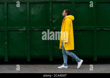 Un homme sous un imperméable jaune descend dans la rue par temps de pluie à côté d'un conteneur vert, vue latérale. Extérieur. Banque D'Images