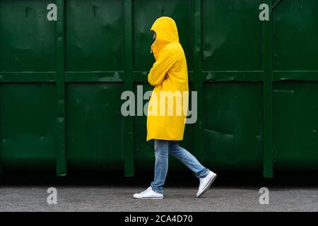 Un homme sous un imperméable jaune descend dans la rue par temps de pluie à côté d'un conteneur vert, vue latérale. Extérieur. Banque D'Images