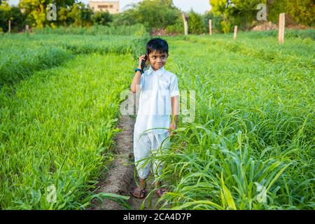 Portrait sur enfant indien mignon parlant sur téléphone mobile debout dans l'agriculture champ vert, la technologie dans l'Inde rurale concept. Banque D'Images