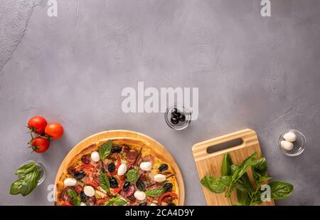 Pizza italienne avec jambon, salami, olives noires, fromage mozzarella, tomates rouges et feuilles de basilic sur fond gris texturé entouré de quelques ingrédients Banque D'Images