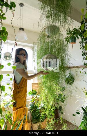 Joyeuse jeune femme jardinière dans des combinaisons orange debout sur un escabeau, touche la boule disco pendante. Verdure à la maison. Amour des plantes. Jardin intérieur Banque D'Images