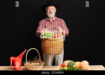 Photo studio de fermier aux cheveux gris avec barbe, debout avec un grand panier en osier rempli de laitue et de brocoli dans les mains, présentant des légumes et des œufs Banque D'Images