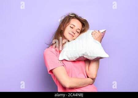 Prise de vue en intérieur d'une jeune femme souriante attrayante avec des plumes dans ses cheveux blond foncé désordonnés, tient l'oreiller, essaie de dormir un peu plus, ne veut pas obtenir Banque D'Images