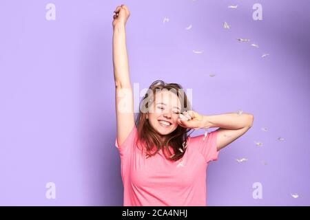 Vue de face d'une jolie fille dansant sous des plumes volantes avec des yeux fermés pendant la fête de combat d'oreillers avec ses amis, se sentant ludique et joyeuse em Banque D'Images