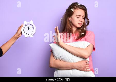 Une fille étudiante fatiguée ne veut pas regarder le réveil, a l'expression somnolente, le regard sombre, a les yeux fermés, la fatigue après la partie tient l'oreiller, a feathe Banque D'Images