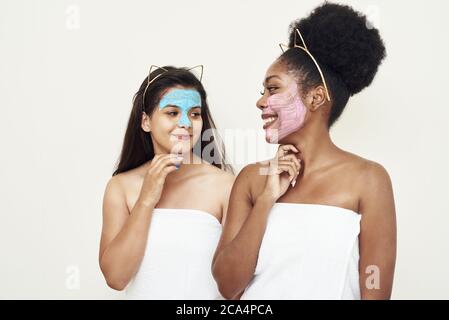 Des jeunes femmes heureux et différentes se tiennent dans le studio dans une serviette et rire, parler entre elles dans des masques cosmétiques. Le concept de l'amitié, de la beauté, des soins personnels et des cosmétiques. Banque D'Images