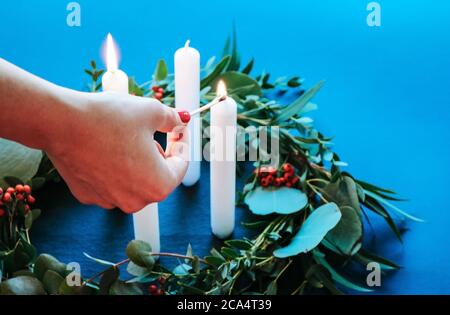 Gros plan sur une femme qui éclaire les bougies de l'Avent sur un décor moderne Couronne de Noël avec feuilles d'eucalyptus isolées sur fond bleu Banque D'Images