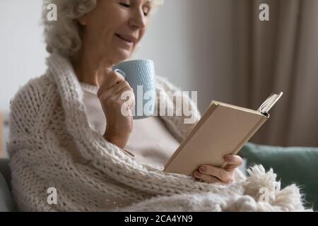 Femme mûre se reposant sur un canapé en train de boire du thé en lisant un livre Banque D'Images