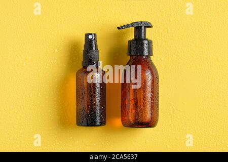 Bouteilles cosmétiques en verre ambré dans des gouttelettes d'eau sur fond jaune. Bouteille d'épice avec crème solaire et bouteille de pompe avec crème solaire. Pose à plat, Banque D'Images