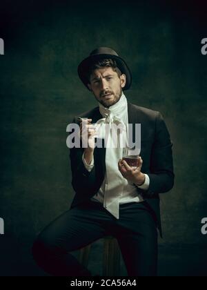 Cigare et brandy. Jeune homme en costume comme Dorian Gray isolé sur fond vert foncé. Style rétro, comparaison du concept ERAS. Beau modèle masculin comme le caractère classique de la littérature, à l'ancienne. Banque D'Images