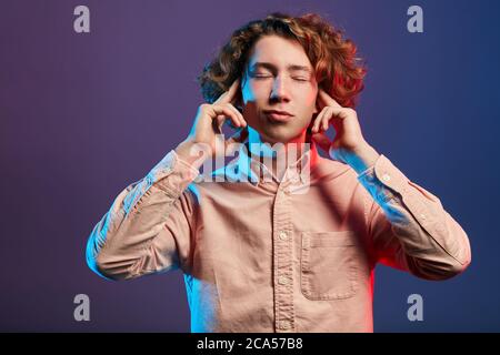 Fatigué redhead jeune personne vêtue dans un décontracté pêche couleur chemise, se reposant dans le club de discothèque, fermer les oreilles avec les mains, trop forte musique, concentré pour da Banque D'Images