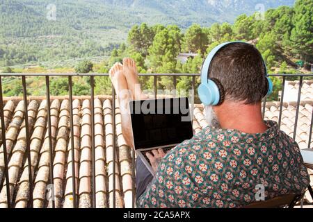 Homme travaillant sur un ordinateur portable avec des écouteurs sur le balcon de sa maison. Travailleur indépendant, travail à distance, technologie, balcon. Restez à la maison. COVID-19 Banque D'Images