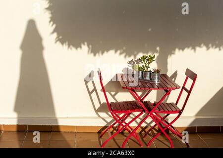 Table et chaises en bois rouge et en métal dans le jardin, à côté d'un mur blanc où vous pouvez voir les ombres d'un arbre et d'un parasol Banque D'Images