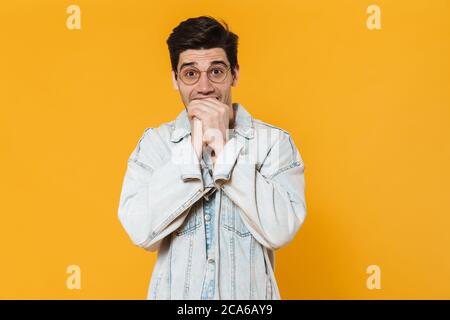 Photo d'un jeune homme effrayé dans des lunettes qui mordant ses doigts et regardant l'appareil photo isolé sur fond jaune Banque D'Images