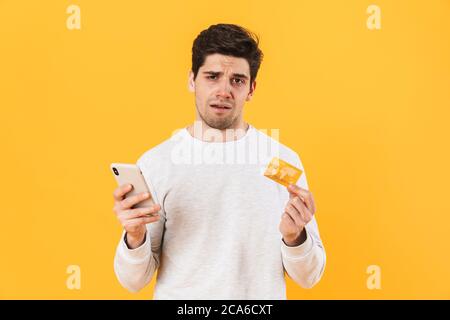 Photo d'un beau malade tenant une carte de crédit et un smartphone isolé sur fond jaune Banque D'Images