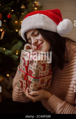 Jeune femme élégante tenant une boîte cadeau de noël et rêvant sous un arbre de noël avec des lumières dans une pièce moderne. Bonne fille dans le chapeau de père noël recevoir le baptême Banque D'Images