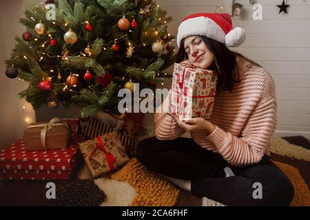 Jeune femme élégante tenant une boîte cadeau de noël et rêvant sous un arbre de noël avec des lumières dans une pièce moderne. Bonne fille dans le chapeau de père noël recevoir le baptême Banque D'Images