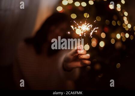 Bonne femme tenant feu d'artifice à l'arbre de noël avec des lumières dorées. Une fille élégante avec un petit feu de bois qui fête dans une pièce sombre de fête. Bonne année Banque D'Images