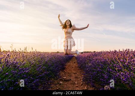 Belle femme en robe sautant dans le champ de lavande fleuri pendant le coucher du soleil. Vue avant Banque D'Images