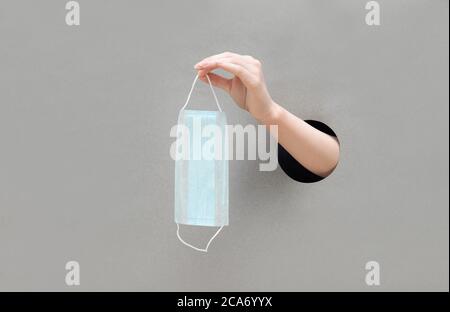 Une main femelle tient un masque médical sur fond gris. Main dans un trou de papier. Prévention du coronavirus, Covid-19. Concept de soins de santé. Banque D'Images
