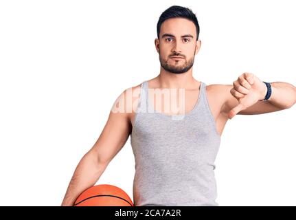 Jeune homme beau tenant le ballon de basket-ball avec le visage en colère, signe négatif montrant le désarroi avec les pouces vers le bas, concept de rejet Banque D'Images