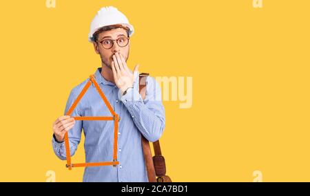 Beau jeune homme avec ours portant architecte hardhat tenant projet de construction couvrant la bouche avec la main, choqué et peur pour erreur. Surpris expre Banque D'Images