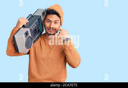 Beau jeune latino-américain tenant une boombox, écoutant de la musique pointant du doigt vers le visage et le nez, souriant gai. Concept de beauté Banque D'Images