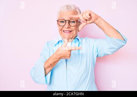 Senior belle femme avec les yeux bleus et les cheveux gris portant des lunettes souriantes faisant cadre avec les mains et les doigts avec le visage heureux. Créativité et photog Banque D'Images