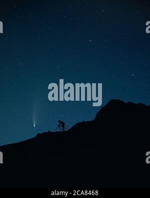 Silhouette d'homme à vélo dans les montagnes sous le ciel nocturne avec des étoiles et une comète de Neolise. Banque D'Images