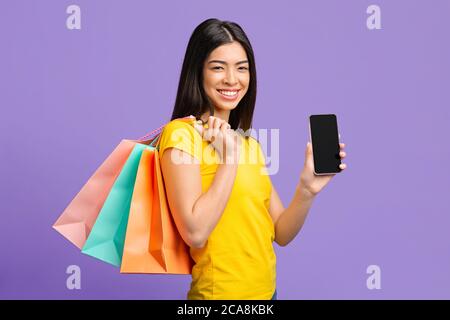 Achats en ligne. Happy Asian Girl avec des cabas-sacs démontrant l'écran noir de smartphone Banque D'Images