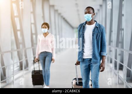 Des Africains heureux en masques médicaux marchant avec des bagages au terminal de l'aéroport Banque D'Images