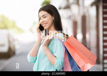 Concept de consumérisme. Asian Girl porter des sacs d'achats et parler sur le téléphone portable à l'extérieur Banque D'Images