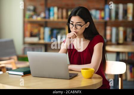 Focalisé asiatique fille étudiant en lunettes étudier en ligne avec ordinateur portable dans le café Banque D'Images