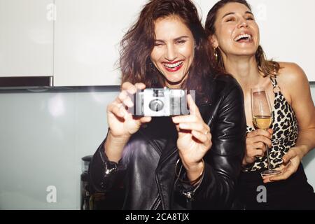 Les femmes gaies font la fête à la maison en buvant du vin. Femme prenant une photo sur un appareil photo jetable lors d'une fête à la maison. Banque D'Images