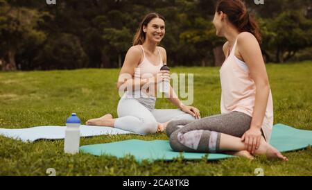 femmes de fitness assises sur des tapis de yoga et se relaxant pendant l'entraînement dans un parc. Souriante femmes amis prendre une pause pendant l'entraînement boire de l'eau. Banque D'Images