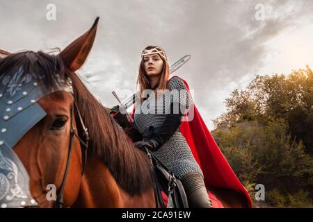 Une belle fille avec une épée dans un costume de fantaisie médiéval. Une femme dans une chaîne de courrier, un manteau, une couronne, sur un cheval en munitions de combat. Une fille à cheval un hors Banque D'Images