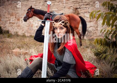 Une fille dans un costume médiéval regarde dans l'appareil photo, se trouve sur l'herbe, un cheval tombe de derrière dans le fond de la forteresse. Une femme avec une épée Banque D'Images