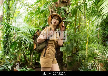 Femme botaniste vêtue de style safari en serre. Naturaliste en vêtements kaki, gants de castor avec sac à dos marche dans une serre entourée de palmiers Banque D'Images