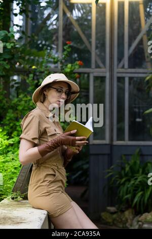 Femme botaniste vêtue de style safari en serre. Naturaliste portant un chapeau et des vêtements kaki, gants de castor se reposant et lisant un livre entouré de Banque D'Images
