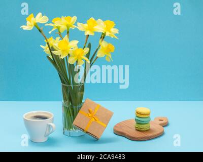 Bouquet de jonquilles jaunes dans un vase en verre, une boîte cadeau, une tasse de café et des macarons sur une planche à découper en bois sur fond bleu Banque D'Images