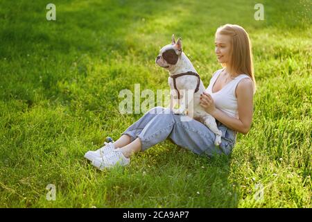 Vue de dessus de la jeune femme heureuse assise sur l'herbe avec le joli boudogue français. Magnifique caucasienne souriante fille appréciant le coucher du soleil d'été, tenant le chien à genoux dans le parc de la ville. Amitié humaine et animale. Banque D'Images