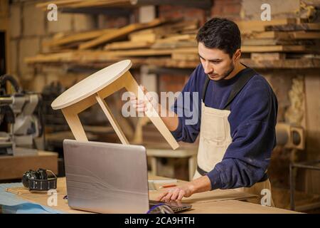 un jeune fabricant de meubles fait habilement une chaise en bois sur un établi et utilise un ordinateur portable tout en travaillant seul dans son grand atelier de travail du bois, l'artisanat et c Banque D'Images