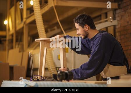 jeune charpentier caucasien mesurant la hauteur de la chaise en atelier, préparant des meubles faits à la main pour les clients Banque D'Images