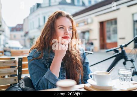 Portrait de triste rouge curled long cheveux caucasien jeune fille assise sur un café confortable terrasse extérieure sur la rue et regardant la rue. Jeune femme t Banque D'Images